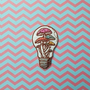 Könnte beinhalten: Ein gestickter Aufnäher mit einer Glühbirne, in der sich ein Büschel bunter Pilze befindet. Die Glühbirne ist weiß mit einem braunen Sockel und die Pilze sind rosa, orange, gelb und blau.