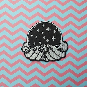 Op de afbeelding: Een geborduurde patch in zwart en wit van twee handen die een kristallen bol vasthouden met zilveren sterren op een zwarte achtergrond.