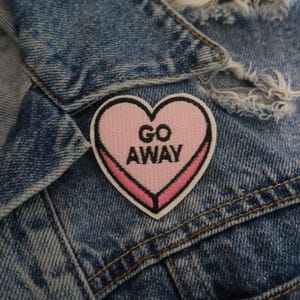 Op de afbeelding: Een roze hartvormige patch met de tekst "GO AWAY" in zwarte letters, genaaid op een blauw denim jack.