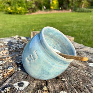 May include: A light blue ceramic salt cellar with the word "SALT" embossed on the side. The salt bowl has a rounded shape and contains a small wooden spoon. The salt cellar sits on a weathered wooden surface.