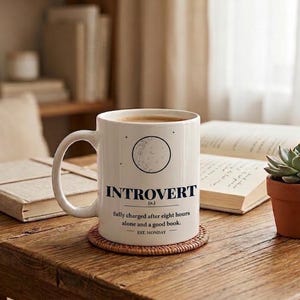 Pode incluir: Uma caneca de cerâmica branca com um gráfico preto de uma lua e o texto: INTROVERT (n.) fully charged after eight hours alone and a good book. EST. MONDAY. A caneca tem uma asa curva e repousa sobre um porta-copos redondo entrançado.