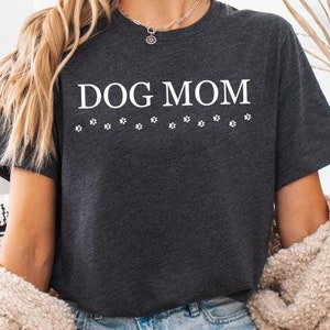 Puede incluir: Una camiseta gris oscuro con el texto "DOG MOM" en letras blancas y una fila de huellas de patas debajo del texto.