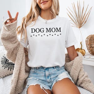 Puede incluir: Camiseta blanca con el texto "DOG MOM" y una fila de huellas de patas debajo. La modelo lleva la camiseta con pantalones cortos de mezclilla y una chaqueta de punto crema.