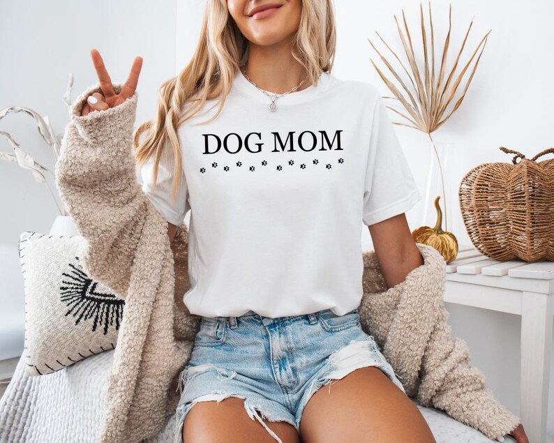 Puede incluir: Camiseta blanca con el texto "DOG MOM" y una fila de huellas de patas debajo. La modelo lleva la camiseta con pantalones cortos de mezclilla y una chaqueta de punto crema.