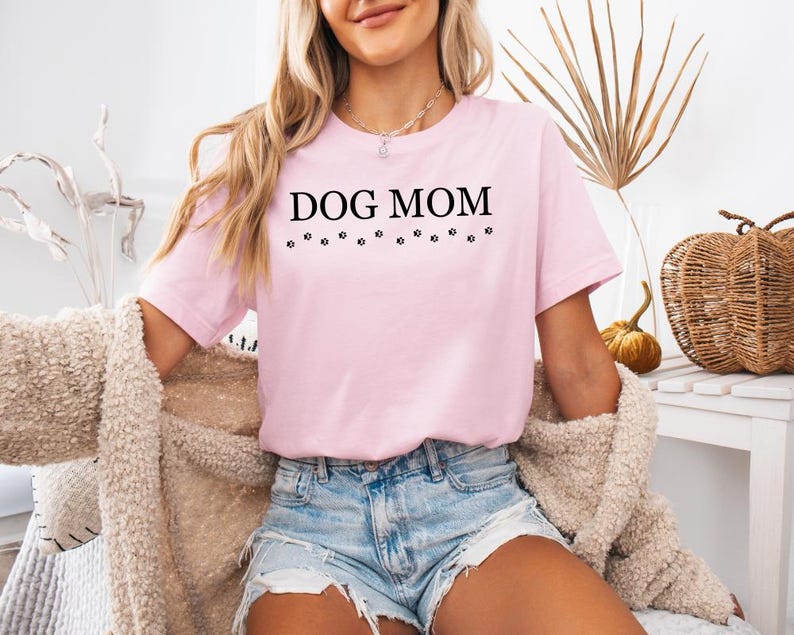 Puede incluir: Una camiseta rosa con el texto "DOG MOM" en letras negras y una fila de huellas de patas debajo del texto.