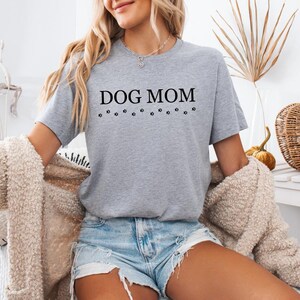 Puede incluir: Una camiseta gris con el texto "DOG MOM" en letras negras y una fila de huellas de patas debajo del texto.