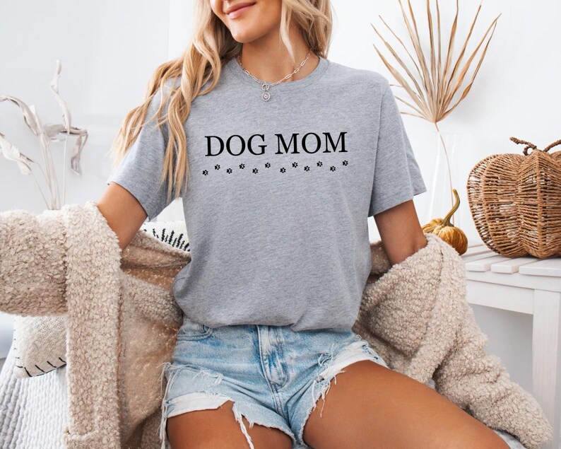 Puede incluir: Una camiseta gris con el texto "DOG MOM" en letras negras y una fila de huellas de patas debajo del texto.