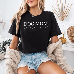 Puede incluir: Una camiseta negra con el texto "DOG MOM" en letras blancas y una fila de huellas de patas debajo del texto.