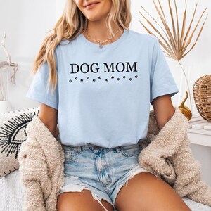 Puede incluir: Una camiseta azul claro con el texto "DOG MOM" en letras negras y una fila de huellas de patas debajo del texto.