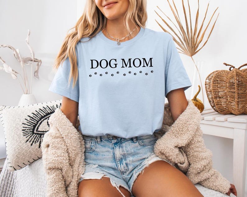Puede incluir: Una camiseta azul claro con el texto "DOG MOM" en letras negras y una fila de huellas de patas debajo del texto.