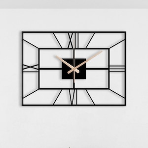 May include: A black metal framed clock with Roman numerals hangs on a white wall above a dining table with four wooden chairs. The table has a wooden top and black metal legs. There is a small plant on the table.
