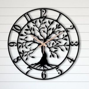 May include: Black metal wall clock featuring a tree of life design. The clock has Roman numerals and a wooden hand. The clock is hanging on a white wall with a wooden bench and red lanterns in front of it.