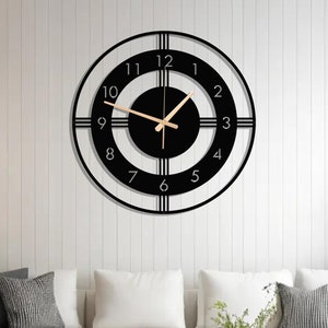 Op de afbeelding: Een zwart-witte klok met een gouden uur- en minutenwijzer. De klok hangt aan een witte muur in een woonkamer. De klok heeft een modern ontwerp met een zwarte lijst en witte cijfers.