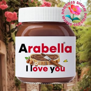 May include: A jar of Arabella hazelnut spread with a white lid. The label features the brand name, an illustration of spread on toast, and the words "I love you". The jar is set against a blurred outdoor background.