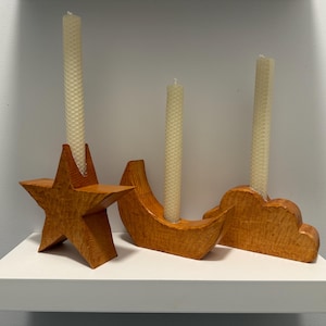 May include: Three wooden candle holders in the shapes of a star, crescent moon, and cloud. Each holder is stained a warm orange color and holds a tall, cream-colored candle. The candle holders are displayed on a white shelf.