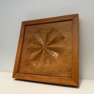 May include: A square, wooden wall art piece with a carved floral design. The artwork is framed in a matching wood and has a warm, natural finish. The carved design is centered on the square.