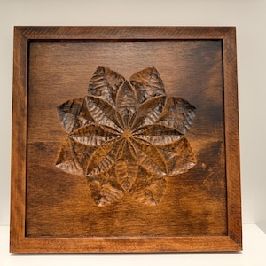 May include: A square, wooden wall art piece with a carved floral design. The artwork features a dark brown frame and a detailed, three-dimensional flower carving in the center. The wood has a rich, warm tone.