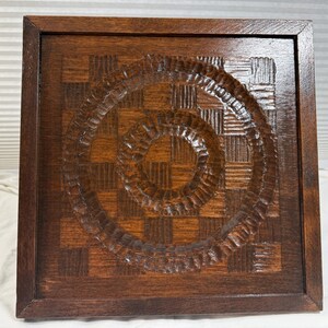 May include: A square wooden wall art piece with a dark brown finish. The artwork features a carved spiral design in the center, surrounded by a checkered pattern. The piece is framed with a dark brown border.