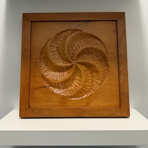 May include: A square, wooden wall art piece with a carved spiral design. The artwork is framed and has a warm, natural wood tone. The spiral is the focal point, with detailed grooves.