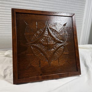 May include: A square, wooden wall art piece with a compass design carved into the surface. The compass features a central starburst pattern and directional arrows. The wood has a dark brown stain and a framed edge.