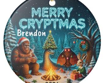 May include: A round ceramic ornament featuring the text MERRY CRYPTMAS, Brendon, and 2026. The illustration depicts Bigfoot, the Loch Ness Monster, Mothman, and a jackalope gathered around a campfire in a snowy forest with Christmas decorations.