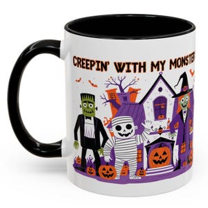 May include: White ceramic Halloween mug with a black handle and rim. The mug features a cartoon illustration of Frankenstein, a mummy, and a witch in front of a haunted house, with the text "CREEPIN' WITH MY MONSTER".