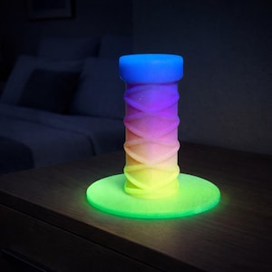 May include: A cylindrical object with a textured, crisscross pattern that transitions in color from blue at the top to purple, pink, orange, and yellow at the base. The object stands upright on a circular base that glows with a bright green light.