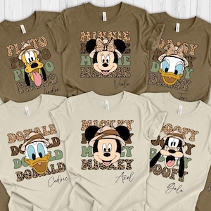 May include: A set of ten brown and beige t-shirts with a safari theme. Each shirt features a different Disney character wearing a safari hat and surrounded by a leopard print pattern. The characters include Mickey Mouse, Minnie Mouse, Donald Duck, Daisy Duck, Goofy, and Pluto. The shirts have names printed on the bottom.