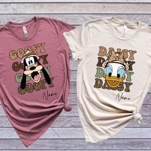 May include: Two t-shirts with a safari theme. One features Goofy with the text "Goofy Goofy Goofy Goofy" and the other features Daisy Duck with the text "Daisy Daisy Daisy Daisy". Both shirts have a brown and green leopard print design.