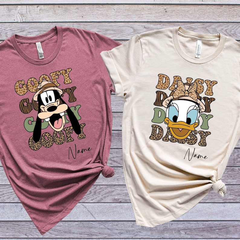 May include: Two t-shirts with a safari theme. One features Goofy with the text "Goofy Goofy Goofy Goofy" and the other features Daisy Duck with the text "Daisy Daisy Daisy Daisy". Both shirts have a brown and green leopard print design.
