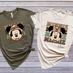 May include: Two t-shirts with a jungle theme. The green shirt features Mickey Mouse wearing a safari hat and the text "Mickey Mickey Mickey". The beige shirt features Minnie Mouse wearing a safari hat and the text "Minnie Minnie Minnie". Both shirts have a leopard print design.