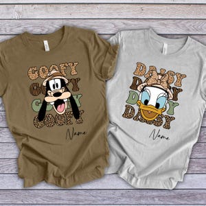 May include: Two brown and grey t-shirts with a safari theme. The brown shirt features Goofy with the text "Goofy Goofy Goofy Goofy" and the grey shirt features Daisy Duck with the text "Daisy Daisy Daisy Daisy".