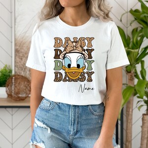 May include: White t-shirt with a cartoon duck wearing a bow and a leopard print design. The text "Daisy Daisy Daisy" is printed on the shirt with the name "Name" below it.