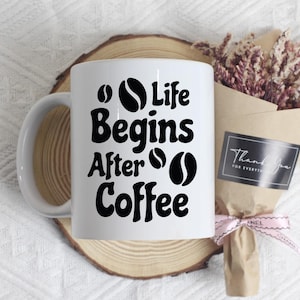 Könnte beinhalten: Weiße Kaffeetasse mit dem schwarzen Text "Life Begins After Coffee" und Kaffeebohnen-Grafiken. Auf einem Holzuntersetzer befindet sich die Tasse. Ein Strauß Trockenblumen in braunem Papier ist ebenfalls zu sehen.