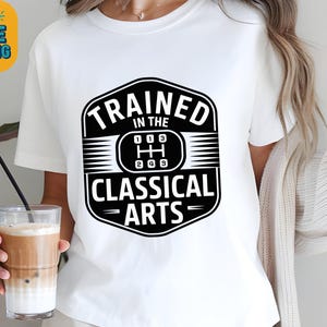 Könnte beinhalten: Weißes T-Shirt mit einer schwarz-weißen Grafik. Die Grafik zeigt "TRAINED IN THE CLASSICAL ARTS" mit einem Schaltdesign. Das Shirt ist ein lässiger Stil, geeignet für den Alltag.