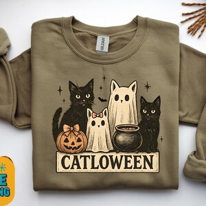 Puede incluir: Sudadera verde oliva con un diseño de Halloween. El diseño incluye gatos negros, una calabaza, un caldero y un fantasma, con la palabra "CATLOWEEN" debajo. Una prenda cómoda y festiva.