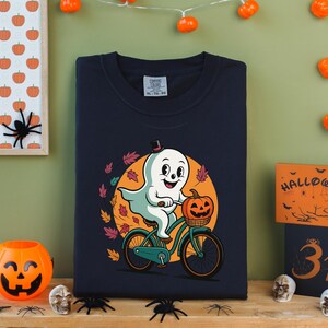 May include: Navy blue sweatshirt featuring a cartoon ghost riding a bicycle with a pumpkin basket. The ghost wears a top hat and is surrounded by autumn leaves. Halloween-themed decor includes a pumpkin bucket, spider decorations, and a framed pumpkin print.