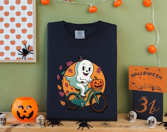 Könnte beinhalten: Marineblaues Sweatshirt mit einem Cartoon-Geist, der mit einem Kürbis-Korb Fahrrad fährt. Der Geist trägt einen Zylinder und ist von Herbstblättern umgeben. Halloween-Dekorationen umfassen einen Kürbis-Eimer, Spinnen und einen gerahmten Kürbis-Druck.