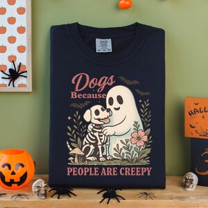 May include: Navy blue t-shirt with a graphic of a ghost hugging a dog skeleton, surrounded by flowers and the text "Dogs Because People Are Creepy." The shirt is part of a Halloween-themed collection.