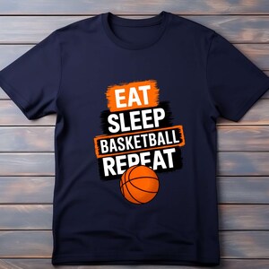 Könnte beinhalten: Ein marineblaues T-Shirt mit dem Text "Eat Sleep Basketball Repeat" in Weiß und Orange. Ein Basketball befindet sich mittig unter dem Text.