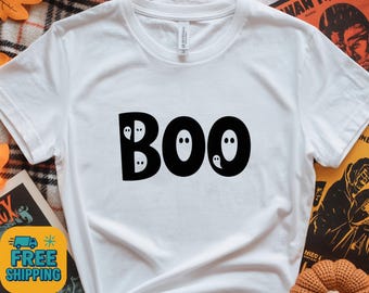 May include: White t-shirt featuring the word "BOO" in bold, black letters. The "O"s are designed with small ghost faces. A playful Halloween-themed design.