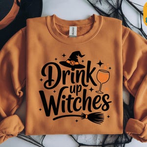 Puede incluir: Una sudadera naranja con la frase "Drink up Witches" en negro, con un sombrero de bruja, una copa de vino y una escoba. Ideal para Halloween o un evento temático de brujas.