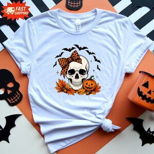 Könnte beinhalten: Ein weißes Kurzarm-T-Shirt mit einer Grafik eines menschlichen Schädels, der eine Schleife mit Leopardenmuster trägt, platziert über orangefarbenen Herbstblättern, Eicheln und einem Kürbis, mit einer Gruppe schwarzer Fledermäuse darüber.