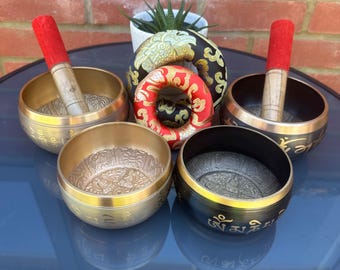 Peut inclure: Plusieurs bols chantants tibétains dorés et noirs, ornés de motifs et d'inscriptions complexes. L'image présente des maillets rouges et en bois, une petite plante succulente et des coussins décoratifs. Les bols sont de différentes tailles.