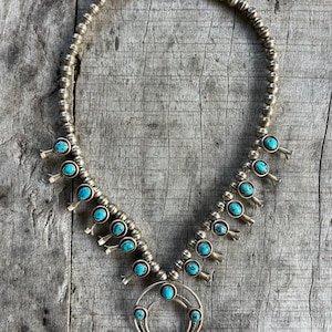 Puede incluir: Collar de plata estilo squash blossom con piedras turquesas. El collar presenta una cadena de cuentas y un colgante en forma de media luna con múltiples detalles turquesas. Las piedras turquesas son de color azul vibrante.