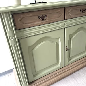 May include: A sage green and brown painted wooden cabinet with two drawers and two doors. The cabinet has ornate details, including decorative drawer pulls and door panels. Two black candle holders and a white vase are on top.