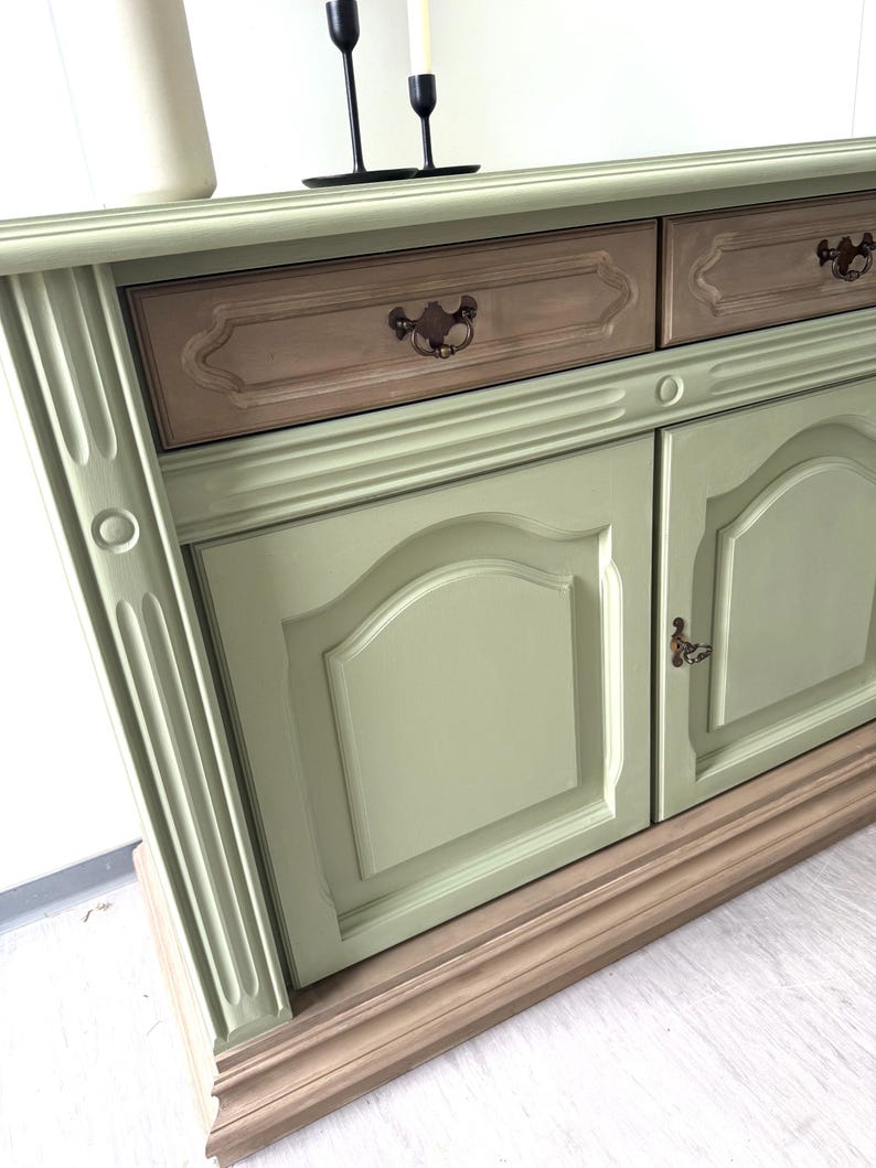 May include: A sage green and brown painted wooden cabinet with two drawers and two doors. The cabinet has ornate details, including decorative drawer pulls and door panels. Two black candle holders and a white vase are on top.