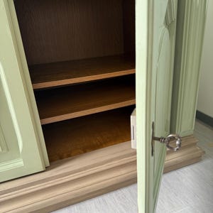 May include: A light green cabinet with two doors, one slightly ajar, revealing two wooden shelves. The cabinet has a decorative base and a key in the lock. The interior is a warm brown.
