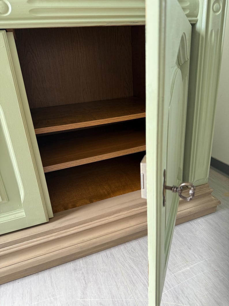 May include: A light green cabinet with two doors, one slightly ajar, revealing two wooden shelves. The cabinet has a decorative base and a key in the lock. The interior is a warm brown.