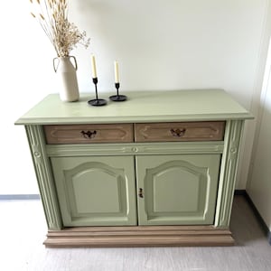 May include: A sage green cabinet with two drawers and two doors. The drawers are a natural wood color with decorative pulls. The cabinet has a light green top and base, with a vase of dried flowers and two black candle holders with lit candles.